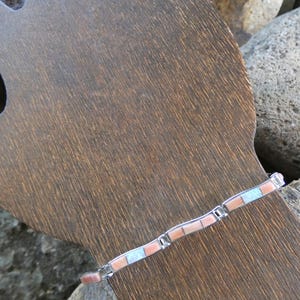 Könnte beinhalten: Ein silbernes Armband mit rechteckigen Einlagen aus Koralle und Türkis. Das Armband wird auf einer Holzform mit dunkler brauner Oberfläche präsentiert. Der Hintergrund zeigt Natursteine.