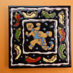 May include: Square ceramic tile with a black background and a white border. The tile features a central figure in yellow and blue, surrounded by colorful chili peppers. Decorative art.