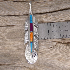 May include: A silver feather pendant with turquoise, purple, orange, and blue inlay. The pendant is about 1 inch long and is shown next to a ruler.