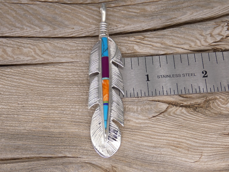 May include: A silver feather pendant with turquoise, purple, orange, and blue inlay. The pendant is about 1 inch long and is shown next to a ruler.