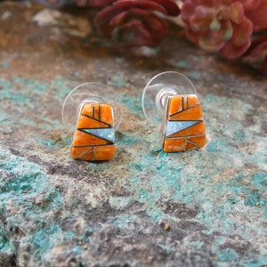 May include: A pair of sterling silver earrings with orange and white inlay. The earrings are in the shape of a triangle and have a geometric design.