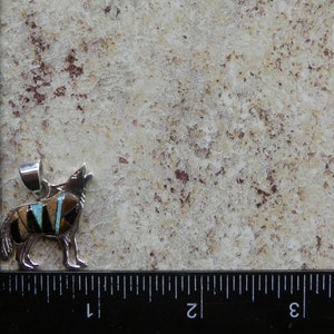May include: Sterling silver pendant featuring a howling wolf design inlaid with turquoise, black, and brown stones. The pendant is approximately 1 inch in length.