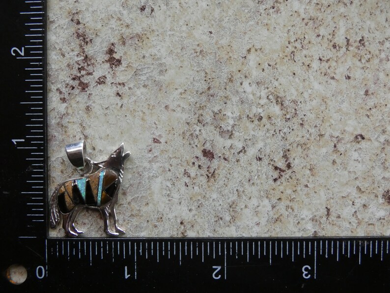 May include: Sterling silver pendant featuring a howling wolf design inlaid with turquoise, black, and brown stones. The pendant is approximately 1 inch in length.