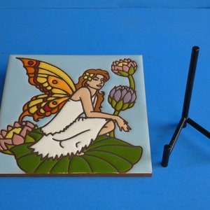 May include: A ceramic tile with a colorful illustration of a fairy with wings sitting on a lily pad. The fairy is wearing a white dress and has brown hair. The tile is on a black metal stand.