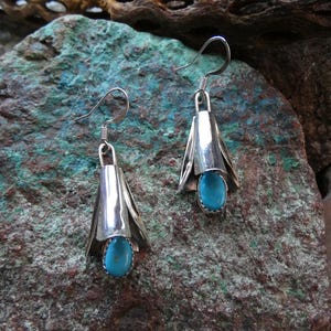 Puede incluir: Un par de pendientes de plata con una piedra turquesa. Los pendientes tienen forma de campana con cierre de gancho. Las piedras turquesas ovaladas están engastadas en plata. Se muestran sobre una superficie rocosa texturizada.
