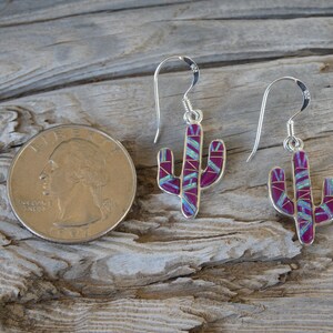 May include: Silver cactus earrings with a magenta and turquoise inlay design. The earrings are shown next to a quarter for size comparison. The earrings have a hook closure.