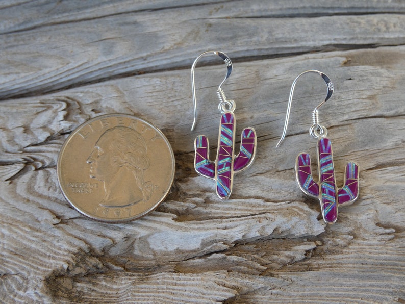 May include: Silver cactus earrings with a magenta and turquoise inlay design. The earrings are shown next to a quarter for size comparison. The earrings have a hook closure.