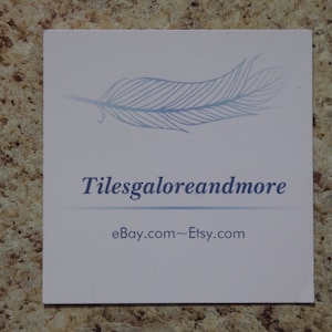 May include: A square, white card with the text "Tilesgaloreandmore" in blue, along with a feather graphic. The card also displays "ebay.com~Etsy.com" in a smaller font.