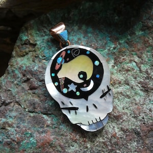 May include: A skull-shaped pendant with a celestial design. The pendant features a mother-of-pearl skull with a copper and turquoise bail. The skull's interior showcases a black background with a yellow shape, stars, and a crescent moon.