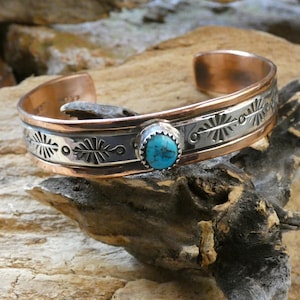 May include: A cuff bracelet featuring a central oval turquoise stone set in a silver bezel. The band is constructed with a copper-colored metal edge and a silver-colored center strip decorated with stamped radial patterns along the outer surface.