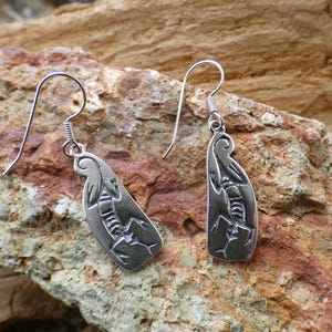 May include: A pair of silver earrings with a lizard design. The earrings are hanging from a silver hook.