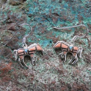 May include: A pair of silver horse earrings with a Southwestern design. The horses have a peach-colored body with black and turquoise accents. The earrings are displayed on a textured, green and brown surface.