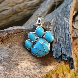 May include: A silver-toned pendant in the shape of a paw print, inlaid with turquoise stones. The paw print design features five rounded turquoise stones for the pads and a larger, intricately designed turquoise stone for the paw's center.