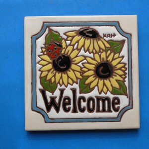 May include: Square ceramic tile with a floral design featuring sunflowers and a ladybug. The word "Welcome" is written in bold, dark brown letters. The tile has a cream-colored background with a blue border.