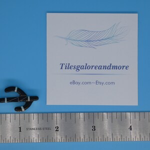 May include: Black pendant with turquoise and silver accents, shaped like an anchor. The pendant is next to a ruler and a card with the text "Tilesgaloreandmore" and "eBay.com~Etsy.com". The background is a solid light blue.