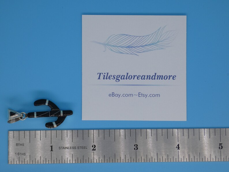 May include: Black pendant with turquoise and silver accents, shaped like an anchor. The pendant is next to a ruler and a card with the text "Tilesgaloreandmore" and "eBay.com~Etsy.com". The background is a solid light blue.