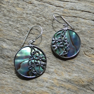 Op de afbeelding: Een paar zilveren oorbellen met een rond ontwerp van abalone-schelp. De schelp heeft een groene, blauwe en paarse iriserende glans. Het zilver is ingewikkeld ontworpen met een bloemmotief.