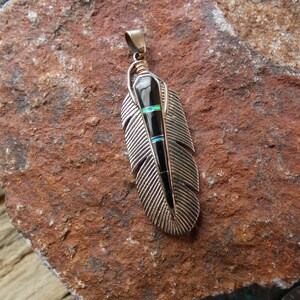 May include: A silver-toned feather pendant with a black center and inlaid turquoise and opal accents. The pendant has a gold-colored bail and is displayed on a textured, reddish-brown surface.