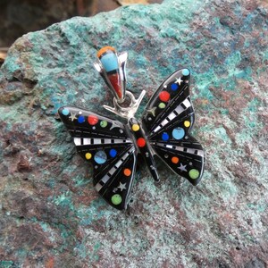 May include: A black butterfly pendant with colorful inlaid details, including red, blue, yellow, and green accents. The pendant has a silver bail and is set against a textured, green and brown stone background.