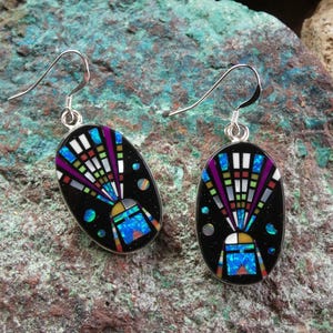 May include: A pair of silver-tone earrings with an oval, black inlay design. The inlay features a colorful geometric pattern with blue, purple, white, and green accents. The earrings have a hook closure and are set against a textured stone background.