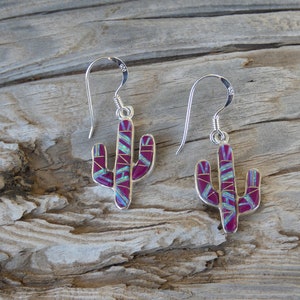 May include: A pair of silver cactus earrings with a magenta and turquoise inlay design. The earrings have a hook closure and are displayed on a wooden surface. The earrings are a southwestern style.