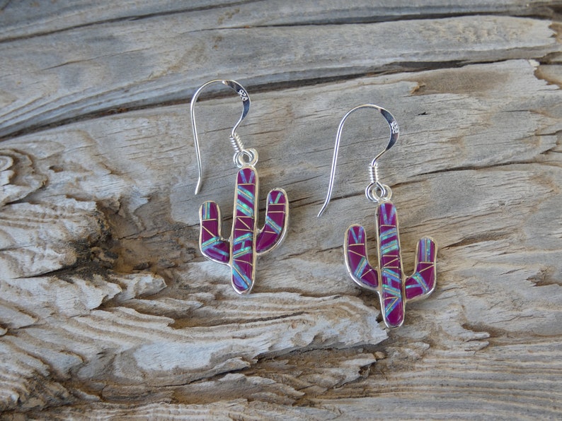 May include: A pair of silver cactus earrings with a magenta and turquoise inlay design. The earrings have a hook closure and are displayed on a wooden surface. The earrings are a southwestern style.