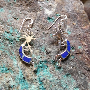 May include: A pair of silver Kokopelli earrings with blue inlay. The earrings feature a stylized figure playing a flute, with a sunburst design above the head. The earrings are displayed on a textured, stone surface.