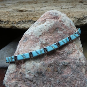 May include: A turquoise and silver bracelet with rectangular turquoise stones and silver links. The bracelet is displayed on a rough, reddish-brown rock, with a blurred background of similar rocks.