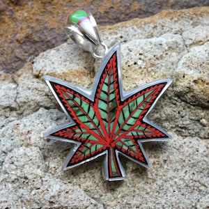 Puede incluir: Un colgante de plata con forma de hoja de marihuana, con incrustaciones rojas y verdes. El colgante tiene un aro para sujetarlo a una cadena.