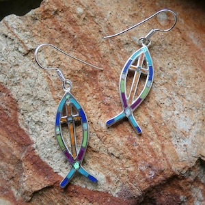 May include: A pair of silver earrings with a colorful mosaic design in the shape of a fish with a cross inside. The earrings have a hook closure.