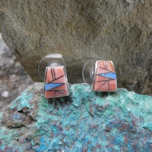 May include: A pair of square stud earrings with a silver frame. The earrings feature a mosaic design with coral, black, and blue inlay. The earrings are set against a natural stone background.