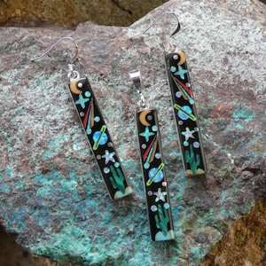 Puede incluir: Un conjunto de pendientes y un colgante negros con un diseño celestial. Las piezas rectangulares presentan una luna creciente, estrellas, planetas y un cactus. Los pendientes tienen ganchos plateados y el colgante tiene una fianza plateada. La joyería se exhibe sobre una superficie rocosa texturizada.