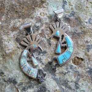 May include: Two silver Kokopelli pendants with turquoise and opal inlay. The pendants are on a rough, gray rock surface.