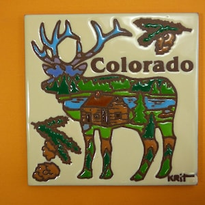 May include: A ceramic tile with a colorful illustration of an elk with antlers, a cabin, and trees. The elk is outlined in brown and filled with green, blue, and brown. The tile has the word "Colorado" written in brown letters.