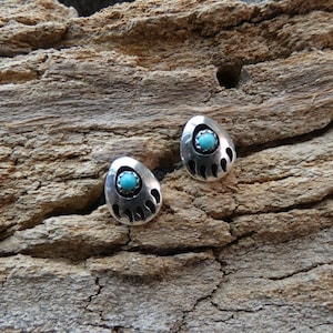 May include: Two silver-toned stud earrings with a turquoise stone center and a bear claw design. The earrings are set against a textured, brown wooden surface. The earrings are oval-shaped.
