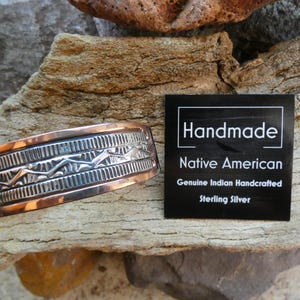 May include: A copper and silver cuff bracelet with a Native American design. The bracelet has a geometric pattern and is next to a black tag that reads "Handmade Native American Genuine Indian Handcrafted Sterling Silver."
