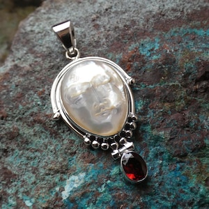 May include: A silver pendant with a white mother-of-pearl face and a red garnet stone hanging from the bottom.