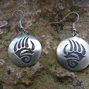 May include: A pair of silver-toned earrings featuring a circular design with a bear paw print. The earrings have a hook closure and are displayed on a textured, mossy surface. The bear paw design is in a darker tone.
