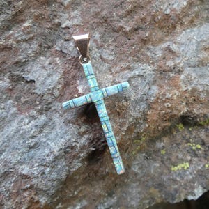 Puede incluir: Un colgante con diseño de cruz con un patrón de mosaico azul claro y blanco. La cruz está suspendida de una fianza dorada. El colgante está sobre un fondo de piedra texturizada gris y marrón.