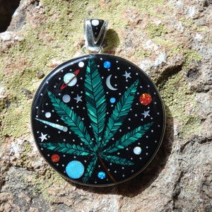 May include: A round silver pendant featuring a green marijuana leaf design. The leaf is set against a black background with white stars, a crescent moon, planets, and comets. The pendant is crafted from inlaid stone.