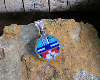 May include: A round pendant with a silver bail. The pendant features a mosaic design with blue, red, white, and yellow elements. The pendant is set against a textured, tan-colored surface.