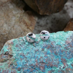 May include: A pair of round stud earrings with a silver setting. Each earring features a white background with black and turquoise inlay designs. The earrings are displayed on a textured, multi-colored rock surface.