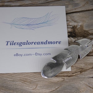 May include: A silver feather charm with a sterling silver stamp. The charm is on a wooden surface with a business card that says "Tilesgaloreandmore eBay.com~Etsy.com".