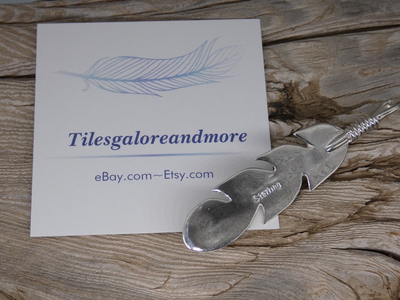 May include: A silver feather charm with a sterling silver stamp. The charm is on a wooden surface with a business card that says "Tilesgaloreandmore eBay.com~Etsy.com".