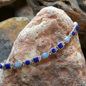 May include: A silver bracelet featuring a series of alternating blue lapis lazuli and oval-shaped opal stones. The bracelet is displayed on a textured, light brown rock surface, showcasing the jewelry's design and color contrast.