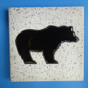 May include: Square ceramic tile featuring a black bear silhouette against a speckled white background. The bear is outlined in a dark brown color. The tile has a rustic, handmade appearance, suitable for home decor.