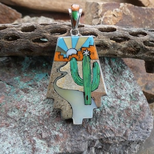 May include: A Southwestern-style pendant with a desert landscape design. The pendant features a turquoise sky with a sun, a green cactus, and a winding road. The pendant is made of various materials and has a silver-colored bail.