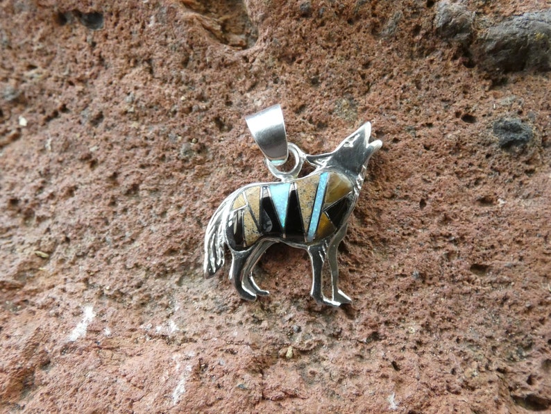 May include: Sterling silver pendant featuring a howling wolf design. The wolf is inlaid with turquoise, black onyx, and tiger's eye stones.