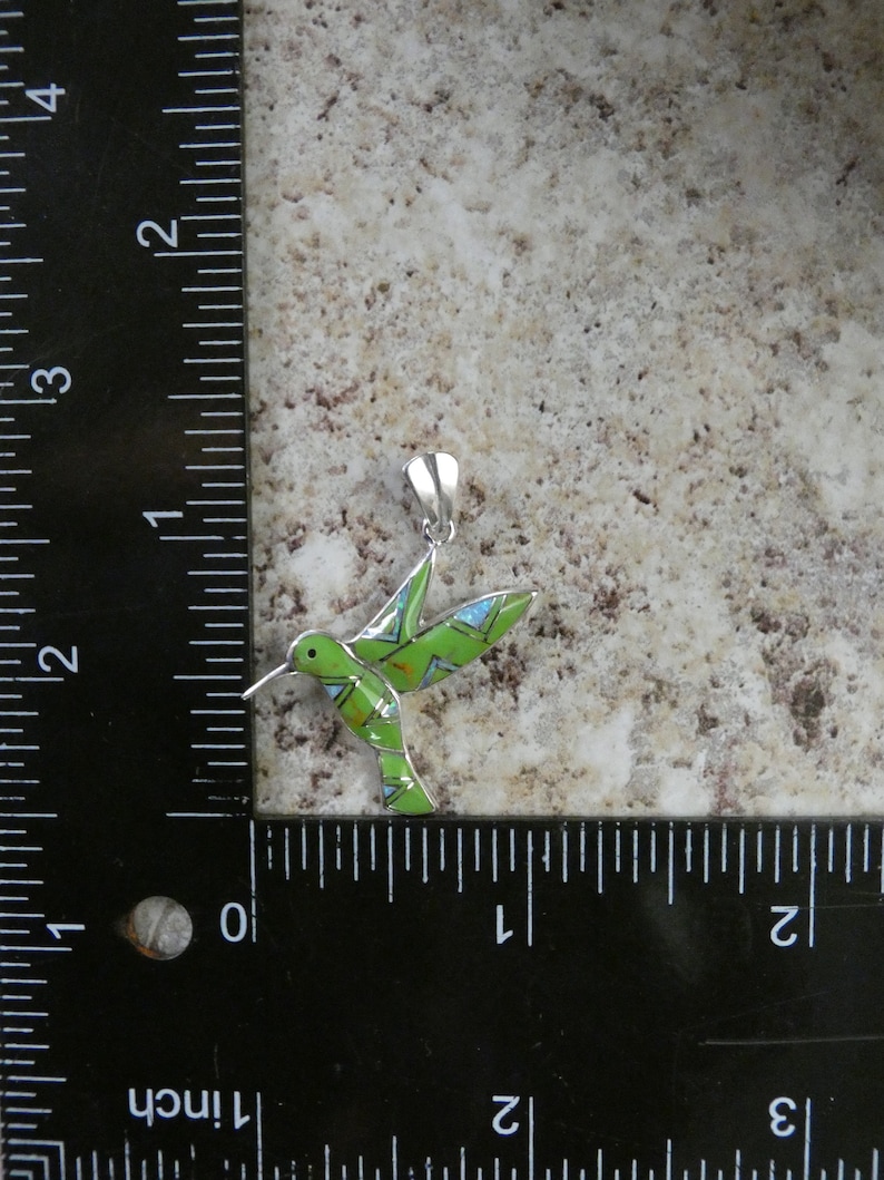 May include: A green and blue hummingbird pendant with silver accents. The pendant is about 1 inch long and is shown next to a ruler.