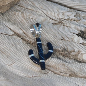 May include: A black cactus pendant with silver accents and a silver bail. The pendant has white and blue details. The pendant is displayed on a weathered wooden surface.